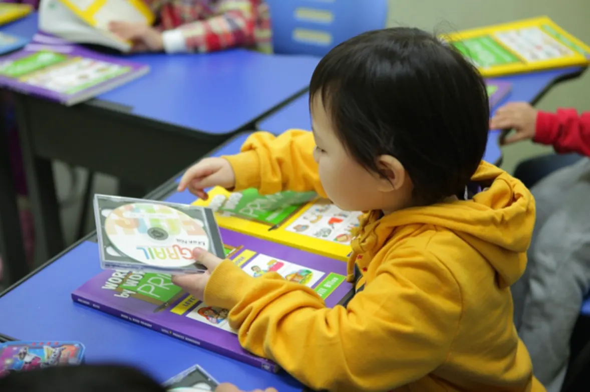 A young student holding a GRAIL phonics CD over a phonics workbook