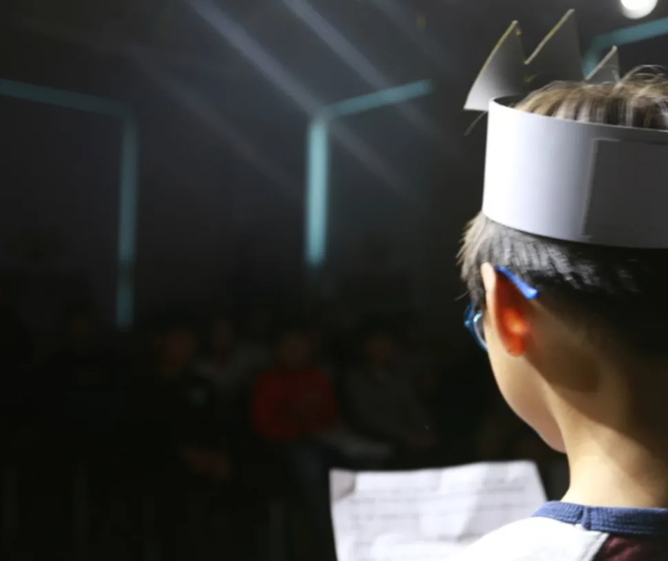 A student in a paper crown reading a script on stage during Project Day