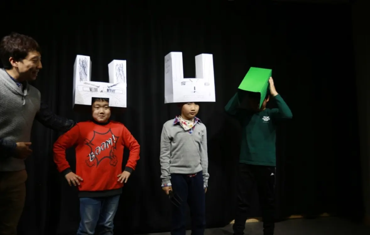 Students wearing handmade paper hats performing on a stage with a teacher